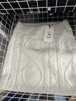 Vit stickad kortkjol med kabelmönster - Supersöt vit stickad kortkjol med bred resår i midjan och snyggt kabelstickat mönster. Kjolen har en mjuk och mysig känsla, perfekt för att skapa en trendig och bekväm look. Passar perfekt till höst och vinter outfits.
