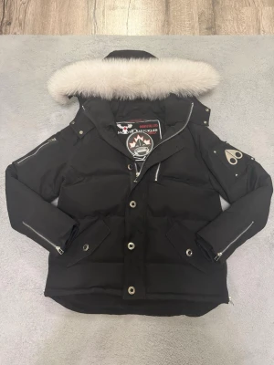 Svart dunjacka Moose Knuckles - Säljer en svart dunjacka från Moose Knuckles med avtagbar huva och vit fuskpäls. Jackan har silverfärgade dragkedjor, tryckknappar och logotyp på ärmen. Flera fickor och detaljer i metall ger en cool look. Perfekt för kalla vinterdagar.