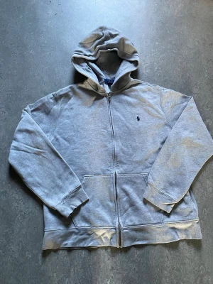 Grå zip hoodie från Polo Ralph Lauren - Snygg grå hoodie med dragkedja från Polo Ralph Lauren. Klassisk design med huva, känguruficka och mörkblå broderad logga på bröstet. Tillverkad i mjuk bomullsblandning som är perfekt för chill dagar. Enkel att matcha med jeans eller sweatpants. Bara att höra av sig vid frågor!
