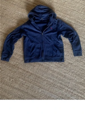 Mörkblå hoodie från Polo Ralph Lauren - Mörkblå hoodie med dragkedja från Polo Ralph Lauren. Klassisk design med huva, kängurufickor och rött broderat logotyp på bröstet. Mjuk bomullsblandning som är skön att ha på sig. jag vill bli av med den pris ej hugget i sten. den är ganska sliten men det är inte så stora defekter, Inga hål. Från axel till midjan är måttet 60cm (se bild 4). Från axel till hand är måttet 60cm (se bild 5). Den är i storlek XL för barn, men den är ganska kort ( kontrollera måtten). DM om ni har några funderingar.