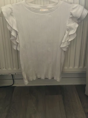 Vit ribbad topp med volangärmar - Supersöt vit topp från Gina Tricot med ribbad struktur och volangdetaljer på ärmarna. Rund halsringning och rak passform. Perfekt för dig som gillar en enkel men ändå trendig look.