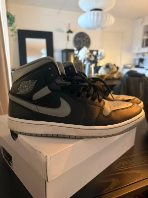 Svarta och gråa Air Jordan 1 sneakers - Säljer ett par klassiska Air Jordan 1 sneakers i svart och grått skinn med ikoniska vingar-loggan på sidan och grå swoosh. Skorna har snörning och vit sula med grå yttersula. Perfekta för dig som vill ha en tidlös och streetinspirerad look.