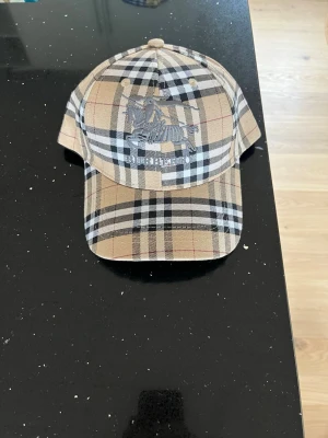 Burberry beige rutig keps - Säljer en klassisk Burberry keps i beige med det ikoniska rutmönstret och broderad logga framtill. Justerbart spänne bak i guld och detaljer med Burberry-tryck på insidan. Perfekt accessoar för dig som vill ha en tidlös och trendig look.