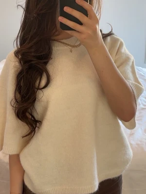 Krämvit stickad tröja med kort ärm - Mysig krämvit stickad tröja med korta ärmar och rund hals. Tröjan har en avslappnad passform och ribbade kanter vid ärmslut och nederkant. Perfekt att styla med jeans eller kjol för en enkel och trendig look.