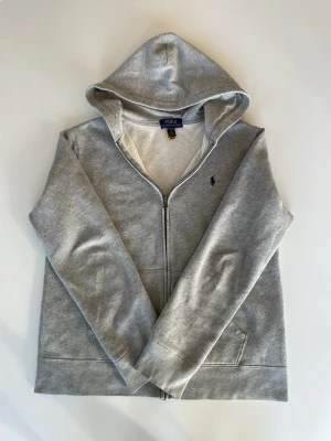 Grå hoodie från Polo Ralph Lauren - Snygg grå hoodie från Polo Ralph Lauren med dragkedja framtill och klassisk huva. Tröjan har två fickor och den ikoniska lilla loggan broderad på bröstet. Tillverkad i mjukt bomullsmaterial som är skönt att bära. Perfekt till jeans eller joggers.