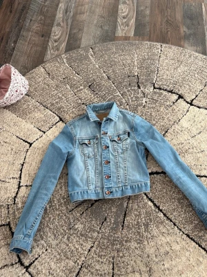 Ljusblå klassisk jeansjacka - Säljer en ljusblå jeansjacka med raka linjer och bruna knappar framtill. Jackan har två bröstfickor med lock och knapp, samt klassisk krage. Perfekt att slänga över en hoodie eller t-shirt för en avslappnad look.