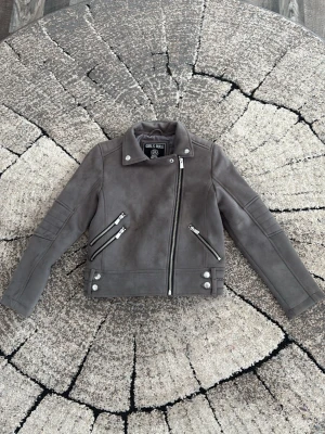 Grå mocka bikerjacka Girls Rule - Grå bikerjacka i mockaimitation från Girls Rule med asymmetrisk dragkedja, tre dragkedjefickor och dekorativa knappar. Jackan har lång ärm och klassisk krage, perfekt för en edgy och trendig stil. Priset kan diskuteras!