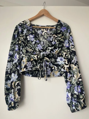 Blommig croppad blus med knyt - Trendig croppad blus med svart botten och blommigt mönster i lila, vitt och grönt. V-ringad med långa ärmar och resår vid ärmslut. Knytning i midjan för justerbar passform. Tunn, mjuk polyester som känns lätt och luftig.