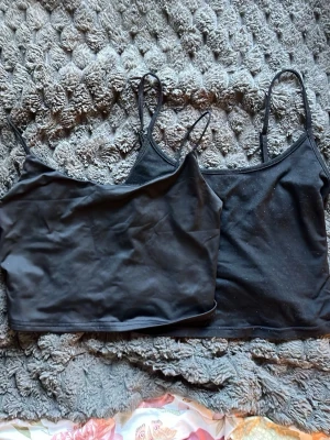 Svarta linnetoppar med smala band - Två svarta linnetoppar med smala axelband och enkel design. Perfekta att matcha med jeans eller kjol för en clean och stilren look. Materialet känns mjukt och stretchigt, vilket gör dem bekväma att bära hela dagen.