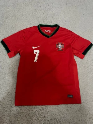 Portugal fotbollströja Nike Ronaldo - Röd Portugal fotbollströja från Nike med nummer 7 och Ronaldo på ryggen. Tröjan har korta ärmar, gröna detaljer vid krage och ärmslut samt Portugals landslagsemblem på bröstet. Tillverkad i lätt och ventilerande material, perfekt för match eller träning.