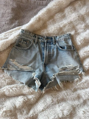 Ljusa högmidjade jeansshorts med slitningar - Säljer ett par ljusblå shorts med mid midja.Shortsen har slitna detaljer framtill, klassiska fem fickor och bälteshällor. Perfekta för sommaren och ger en avslappnad vibe.