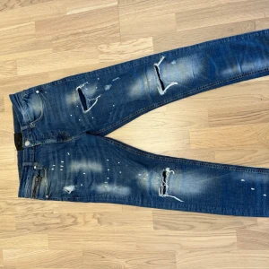 Blå slitna jeans  - Säljer ett par blå jeans med slitningar, hål och vita färgstänk för en riktigt trendig look, Perfekta för dig som gillar streetstyle och vill sticka ut. lite använd och i bra skick. Jag e 170cm och 72kg som ni ser hur den sitter på mig samt finns bild på måtten.