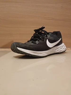 Svarta Nike sneakers med vit sula - Snygga svarta Nike sneakers med vit sula och stor vit Swoosh-logga på sidan. Ovandelen är i mesh och textil för bra andningsförmåga och komfort. Yttersulan har ett greppvänligt mönster, perfekt för vardagsbruk. Klassisk låg modell med snörning.