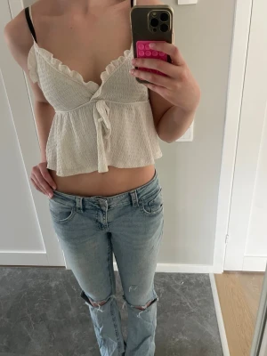 Vit croppad omlottopp med volang - Söt vit croppad topp med omlott-design och volangdetaljer längs halsen. Toppen har knyt framtill och är ärmlös, perfekt för varma dagar. Materialet är lätt och luftigt, vilket gör den bekväm att bära.