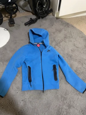 Blå Nike hoodie med dragkedja - Cool blå hoodie från Nike med svart logga på bröstet och svarta detaljer vid dragkedjorna på fickorna. Tröjan har huva och hel dragkedja framtill, perfekt för att slänga på sig när det är lite kyligt. Materialet känns mjukt och sportigt.