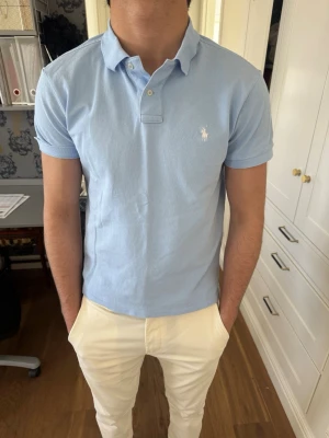 Ljusblå piké från Polo Ralph Lauren - Snygg ljusblå pikétröja från Polo Ralph Lauren i modellen Custom Slim Fit. Tröjan har korta ärmar, klassisk krage och knappar vid halsen. Tillverkad i mjuk bomull och har den ikoniska loggan broderad på bröstet.