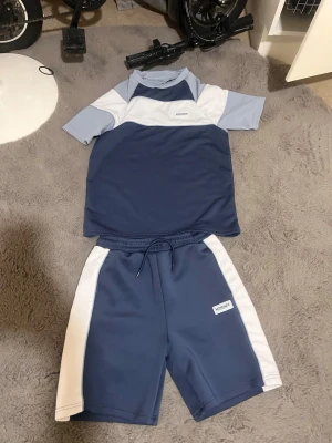 Blått och vitt träningsset meckenzi - Snyggt träningsset från WEEKDAY med matchande t-shirt och shorts i blått och vitt. T-shirten har korta ärmar och rund hals, medan shortsen har dragsko i midjan och vita sidopaneler. Perfekt för gymmet eller sportaktiviteter. Tillverkad i mjukt, funktionellt material.