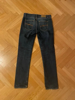 Mörkblå Nudie Jeans Gritty Jackson - Säljer ett par mörkblå slim fit jeans från Nudie Jeans med klassiska bakfickor och orangea sömmar. Jeansen har fem fickor och är tillverkade i denim med snygg tvätt och detaljer. Perfekt för dig som gillar en stilren och modern look.