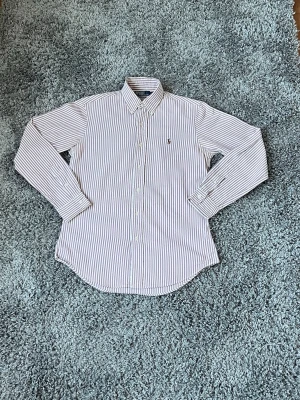 Randig skjorta från Polo Ralph Lauren - Säljer en klassisk långärmad skjorta från Polo Ralph Lauren med vit och blå randigt mönster. Skjortan har knappar framtill och broderad logga på bröstet. Perfekt för dig som gillar stilrena plagg med ikonisk design. Enda defekten är att den är lite gul i armhålan men man tänker inte på det när man har på sig den.