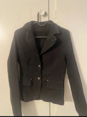 Svart kavaj med knappar - Stilren svart kavaj med två knappar framtill och klassisk krage. Kavajen har två fickor med lock och är figursydd för en snygg passform. Materialet ser ut att vara bomull eller liknande och passar perfekt till olika outfits.