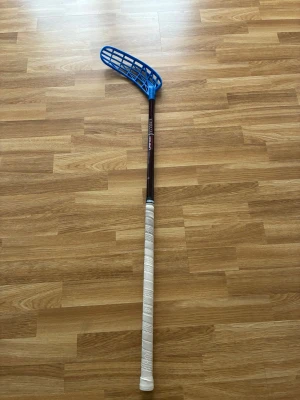 Unihoc  - Säljer en innebandyklubba med svart skaft och blått plastblad. Greppet är lindat med vitt greppband för extra komfort. Perfekt för dig som gillar innebandy och vill ha en lätt och smidig klubba.