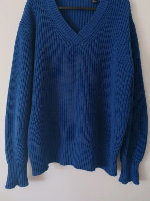 Blå stickad tröja med v-ringning - En blå stickad tröja med djup v-ringning och ribbstickad struktur. Tröjan har långa ärmar och breda muddar vid ärmslut och nederkant. Perfekt för att hålla sig varm och snygg under kyliga dagar.