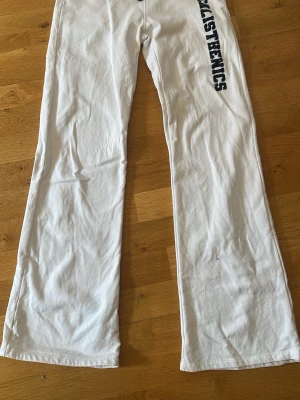 Vita mjukisbyxor från Gina Tricot - Vita mjukisbyxor från Gina med mörkblå tect längs ena benet. Byxorna har raka ben och är låga i midjan. Vet ej varför färgen ser annorlunda ut på andre bilden, längst ner på båda benen finns små fläckar men inget som syns. Säljer så dem är för korta för mig som är 172.🤍