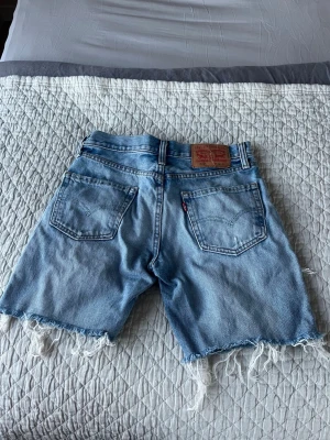 Levi's ljusblå jeansshorts med fransar - Säljer ett par ljusblå jeansshorts från Levi's med råa, fransiga benslut och klassiska bakfickor. Shortsen är grymt snygga med sina slitningar. Perfekta för sommaren och ger en avslappnad vibe.. shortsen är aldrig använda och var för tajta för mig. Storlek 28 i midjan oct köptes för 900 kr på Levis store i Västerås 