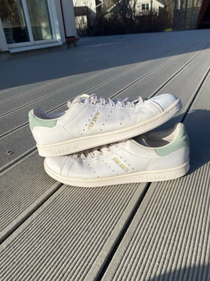 Adidas Stan Smith vita sneakers - Klassiska Adidas Stan Smith sneakers i vitt läder med ljusgröna detaljer på hälen och guldfärgad text på sidan. Skorna har perforerade ränder och en stilren, låg profil. Perfekta för dig som gillar tidlös och clean stil.