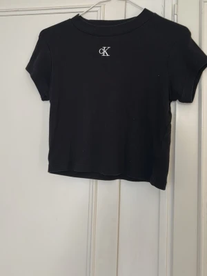 Svart croppad topp från Calvin Klein - Svart croppad topp från Calvin Klein med korta ärmar och ribbad struktur. Diskret CK-logga broderad framtill vid halsen. Perfekt för en stilren och enkel look.