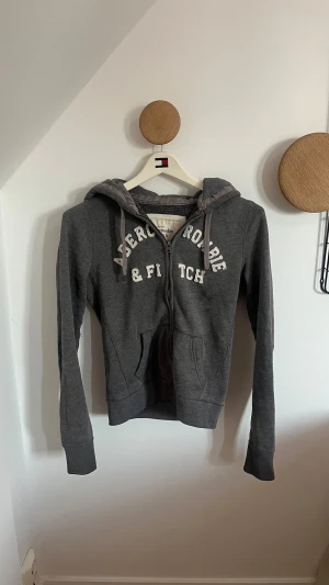 Grå hoodie från Abercrombie & Fitch - Snygg grå hoodie med dragkedja från Abercrombie & Fitch. Stor vit logga på bröstet, två fickor fram och justerbar huva med snören. Perfekt för en chill och avslappnad stil. Mjuk bomullsblandning som känns skön mot huden.
