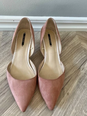 Rosa pumps från Zara kitten heal i mocka  - Snygga pumps i mjuk rosa mocka-look från Zara. Skorna har spetsig tå och låg klack, samt öppen sida för en trendig siluett. Perfekta för dig som vill ha en stilren och modern sko med en feminin touch.