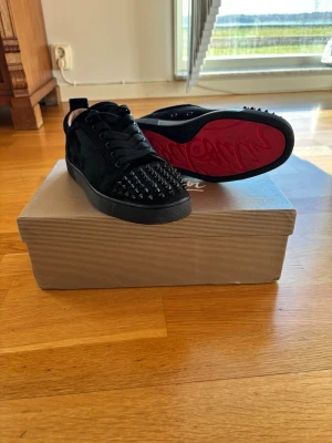 Svarta Louboutin sneakers med nitar - Säljer ett par svarta  Louboutin från med mattsvarta nitar på tån och klassisk röd sula. Skorna har snörning, är låga och tillverkade i mocka och läder. Perfekta för dig som vill sticka ut med en edgy och lyxig stil.