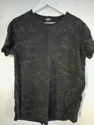 Svart t-shirt med fågelmönster W.BXTR - Svart t-shirt från W.BXTR by William Baxter med diskret fågel- och grenmönster. Klassisk rund halsringning och korta ärmar. Tillverkad i mjuk bomull som känns skön mot huden. Perfekt för dig som gillar subtila detaljer och stilren design.