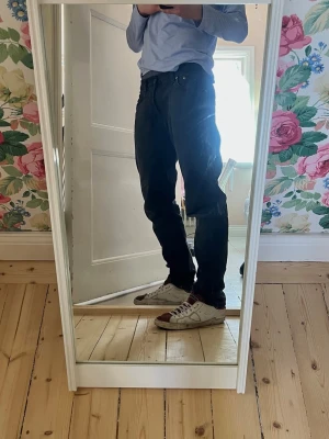 Svarta raka jeans från Nudie - Säljer ett par svarta jeans från Nudie med rak passform och klassisk femficksdesign. Jeansen har normal midja och är tillverkade i ett robust denimtyg som ger en snygg och stilren look. Perfekta för dig som gillar en enkel och clean stil.