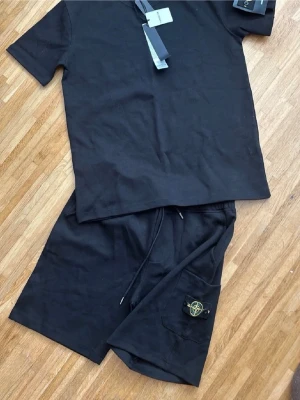 Svart set från Stone Island - Säljer ett svart set från Stone Island med t-shirt och shorts. Setet är gjort i mjukt bomullsmaterial och har en clean look. Shortsen har en ficka med den klassiska Stone Island-loggan i gult och grönt. Perfekt för en avslappnad och stilren outfit.