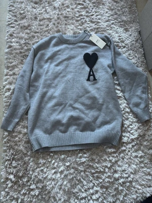 Grå stickad tröja från Ami Paris - Snygg grå stickad tröja från Ami Paris med svart hjärta och bokstaven A broderat på bröstet. Tröjan har rund halsringning, ribbade muddar och nederkant. Perfekt för dig som gillar stilrena detaljer och klassisk design.