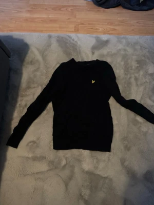 Svart tröja från Lyle & Scott - Svart långärmad tröja från Lyle & Scott med klassisk rund halsringning och den ikoniska gula örnloggan broderad på bröstet. Enkel och stilren design som passar perfekt till jeans eller chinos.