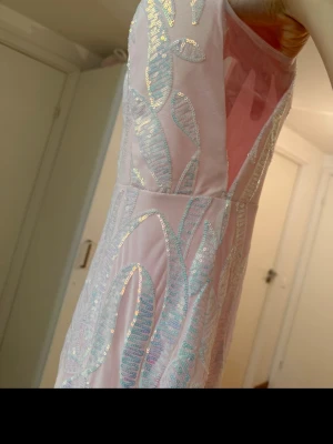 Ljusrosa långklänning med paljetter - Säljer en elegant ljusrosa långklänning med v-ringning och breda axelband. Klänningen har ett skimrande paljettmönster i blått och silver som ger en lyxig känsla. Perfekt för speciella tillfällen där du vill sticka ut.