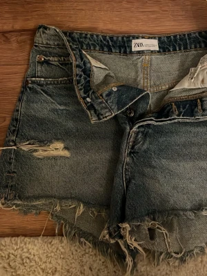 Blå jeansshorts med slitningar från Zara - Säljer ett par blå jeansshorts från Zara. Shortsen har klassisk femficksdesign, knappgylf och fransig nederkant för en avslappnad look. Perfekta för sommaren! Storlek 40!🩷