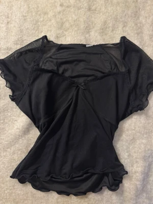 Svart mesh topp med volangdetaljer - Snygg svart mesh topp med korta ärmar och volangdetaljer längs kanterna. Toppen har en lätt transparent look och v-ringad skärning framtill. Perfekt för att addera en edgy touch till din outfit.