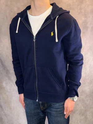Marinblå Ralph Lauren Zip Hoodie - Stl M - Riktigt snygg Ralph Lauren Zip Hoodie i färgen Marinblå med gul pony | Mycket bra skick: 9,2/10, inga defekter | Stl: M, modellen är 177cm och väger 74kg, passar fint | Nypris: Ca 1800kr | Hör av dig vid frågor // Galgen 🫡