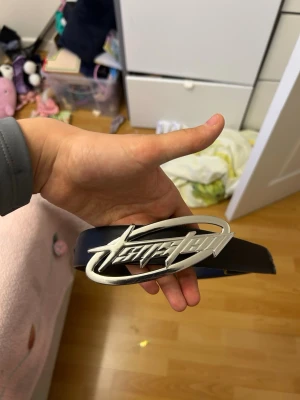 Svart bälte med stor silverfärgad spänne - Coolt svart bälte med ett stort, unikt silverfärgat spänne från Fast Team. Bältet har en klassisk form och är tillverkat i syntetmaterial. Perfekt för att ge din outfit en edgy touch.