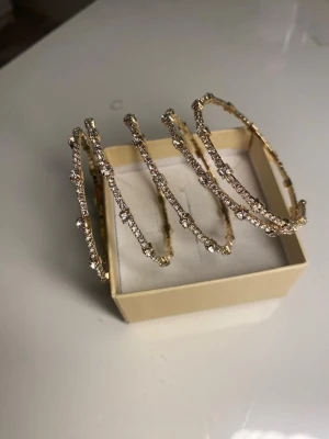 Guldiga armband med strassdetaljer - Säljer ett set med flera tunna armband i guldfärgad metall, dekorerade med gnistrande strass. Armbanden har en rund form och ger en riktigt lyxig och glittrig känsla. Perfekt för dig som vill addera lite extra bling till din outfit.