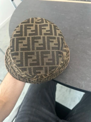 Fendi bucket hat med FF-mönster - Säljer en snygg bucket hat äkta från Fendi i brunt tyg med det klassiska FF-logomönstret i svart. Hatten har en rund form och bred skärm, perfekt för dig som vill sticka ut med en lyxig accessoar. Materialet känns slitstarkt och designen är riktigt trendig.priset kan diskuteras och mer bilder