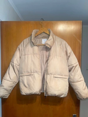 Beige pufferjacka från Lindex - Snygg beige pufferjacka från Lindex med hög krage och dragkedja framtill. Jackan har två stora fickor med dragkedja och är tillverkad i vattenavvisande material. Perfekt för kalla vinterdagar och ger en trendig look.