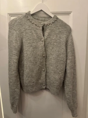 Grå kofta med gnistrande detaljer  - Mjuk lite lurvig grå kofta med rund hals och dekoration längs kragen. Lite noppig. Fina knappar i grått som koftan.