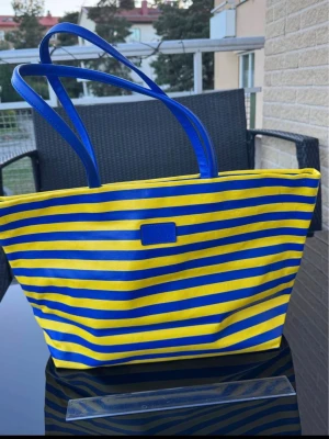 Stor shoppingväska i blått och gult - Ny stor väska med blå och gula ränder. Väskan har två rejäla handtag och en praktisk fack med dragkedja på baksidan. Perfekt för att bära med sig allt man behöver under dagen eller kanske till stranden. Väskan har magnetlås. Skickas eller hämtas på Hägerstensåsen i Stockholm. 🇸🇪