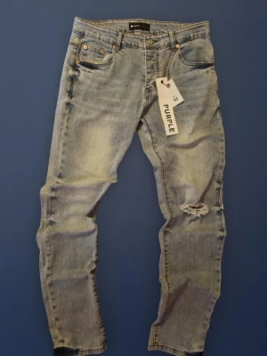 Ljusblå slitna jeans från Purple - Säljer ett par ljusblå jeans från Purple med snyggt slitna detaljer på ena benet. Jeansen har klassisk femficksmodell, normal passform. Perfekta för en avslappnad och trendig look.