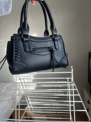 Svart minihandväska med detaljer - Snygg svart minihandväska i konstläder med dubbla handtag och dekorativ dragkedja framtill. Väskan har flätade detaljer på sidorna och en stilren, boxig form. Perfekt accessoar för att lyfta din outfit.